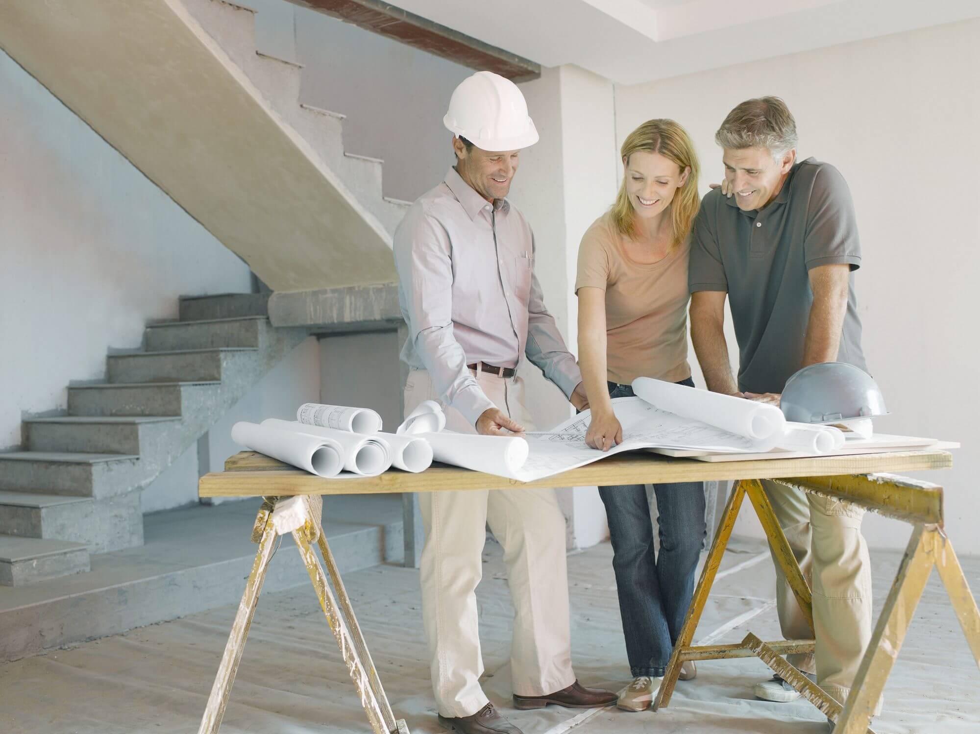 Three professionals discussing construction plans at a table, with rolled blueprints and a hard hat on a worksite, emphasizing collaboration in project planning.