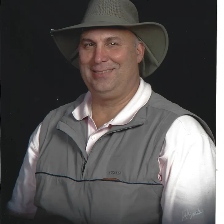 Man smiling, wearing a gray vest and a wide-brimmed hat, against a dark background, representing expertise in industrial refrigeration and consulting services for contractors.