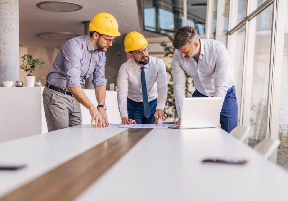 Three professionals in hard hats discussing plans at a conference table with a laptop, emphasizing collaboration and effective communication in project management.